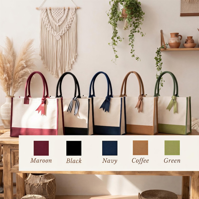 A row of five colorful tote bags with handles, each in a different color, displayed on a wooden table. The bags are arranged in a row, with the first bag being maroon, the second black, the third navy, the fourth coffee, and the fifth green. The background features a wall with a macrame wall hanging and potted plants.