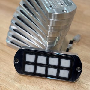May include: A stack of silver rectangular metal housings with rounded edges, and a black control panel with eight illuminated white square buttons. The text "SWITCH-PRO" is visible on the panel.