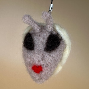 May include: A handmade felt alien face charm with large black eyes, a red heart-shaped mouth, and small antennae. The alien is gray with white accents and is attached to a black lanyard. The charm is a unique, whimsical accessory.