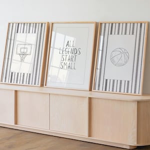 May include: Three framed basketball-themed art prints. Two prints feature a basketball hoop and a basketball on a striped background. The center print has the text "ALL LEGENDS START SMALL" in black. The frames are light wood.