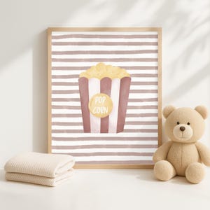 Pode incluir: Impress&atilde;o emoldurada com uma ilustra&ccedil;&atilde;o de pipocas. O recipiente de pipocas &eacute; vermelho e branco com um c&iacute;rculo amarelo que diz "Pop Corn". O fundo tem listras horizontais marrons e brancas. Um urso de pel&uacute;cia e um cobertor dobrado est&atilde;o na imagem.