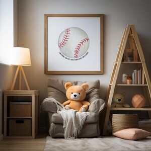 Pode incluir: Uma impressão emoldurada com um baseball em aquarela com costuras vermelhas e o texto "I AM STRONG, I AM FAST, I AM A TEAM PLAYER, I AM A LEADER." Um ursinho de pelúcia está sentado em uma poltrona bege, com um cobertor. Uma estante de madeira e uma mesa lateral também são visíveis.