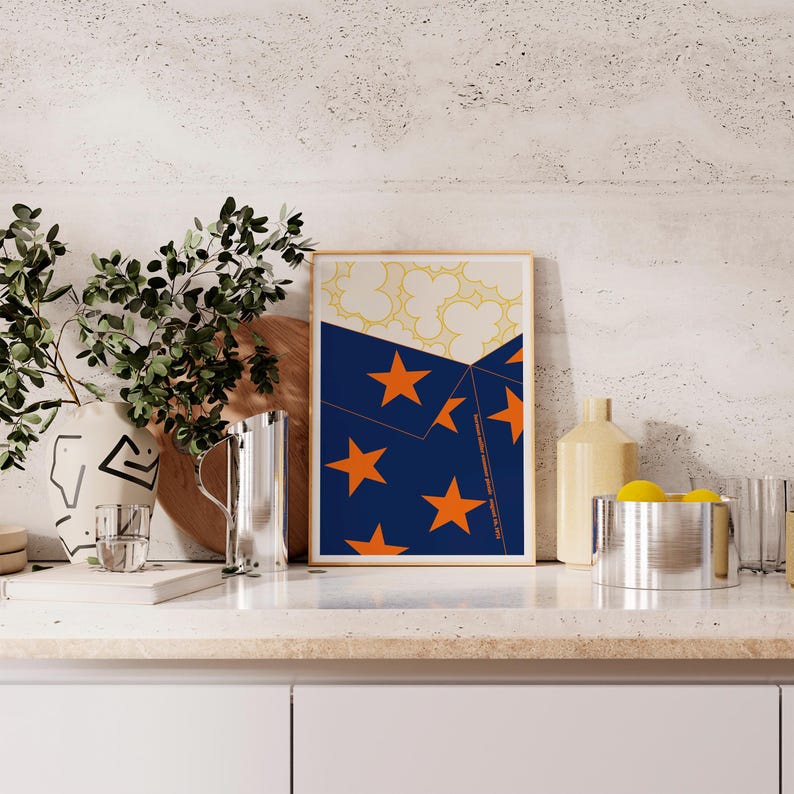 May include: A framed art print with a navy blue section and orange stars, a cream section with yellow floral shapes, and a light brown frame. The print is on a kitchen countertop with other decorative items.