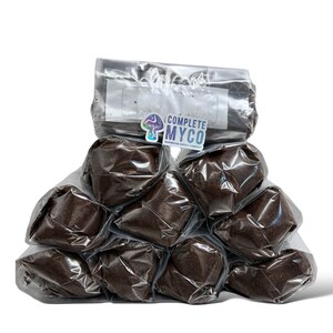 Puede incluir: Una pila de bolsas de plástico selladas llenas de material marrón oscuro. Las bolsas están dispuestas en forma de pirámide. La bolsa superior tiene una etiqueta y una pegatina que dice "COMPLETE MYCO MUSHROOM SUPPLY COMPANY."