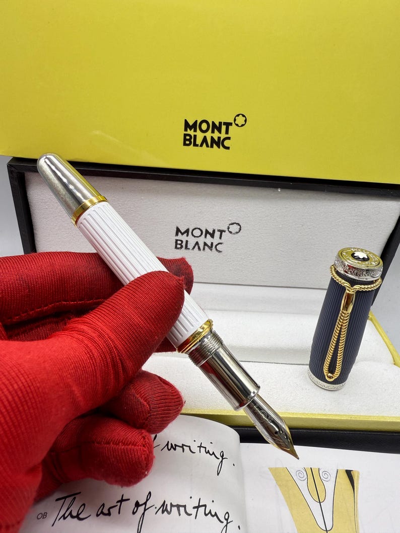 Pu&ograve; includere: Una penna stilografica Montblanc con corpo bianco e dettagli argentati, presentata in una scatola gialla e bianca. La penna ha un pennino dorato ed &egrave; visibile un libretto con la scritta "The art of writing".