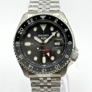 May include: A Seiko 5 Sports automatic GMT watch with a black dial, luminous hour markers, and a red GMT hand. The watch features a stainless steel case and a jubilee-style bracelet, with a black rotating bezel marked with 24-hour numerals.