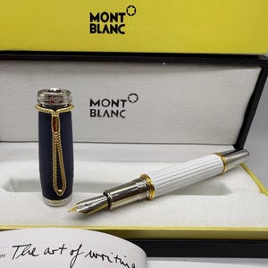 Pu&ograve; includere: Una penna stilografica Montblanc nella sua scatola di presentazione. La penna ha un fusto bianco con dettagli dorati e un pennino argentato. Il cappuccio della penna &egrave; blu navy con dettagli dorati. La scatola &egrave; nera e gialla, con il logo Montblanc.