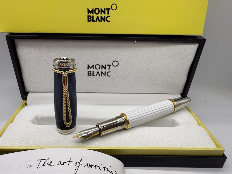 Pu&ograve; includere: Una penna stilografica Montblanc nella sua scatola di presentazione. La penna ha un fusto bianco con dettagli dorati e un pennino argentato. Il cappuccio della penna &egrave; blu navy con dettagli dorati. La scatola &egrave; nera e gialla, con il logo Montblanc.