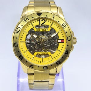 May include: A gold-toned Tommy Hilfiger automatic watch with a yellow face. The watch features a visible inner mechanism, black numerals, and the brand's logo. The watch band is also gold-toned.