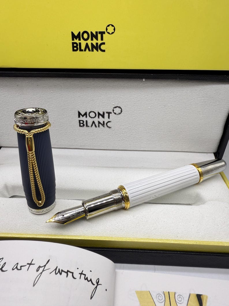 Pu&ograve; includere: Una penna stilografica Montblanc nella sua scatola di presentazione. La penna ha un corpo bianco con dettagli argentati e un pennino dorato. Il cappuccio della penna &egrave; blu navy con dettagli dorati. La scatola &egrave; nera con il logo Montblanc.