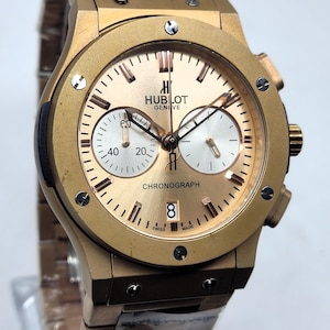 Könnte beinhalten: Eine goldfarbene Hublot Geneve Chronographenuhr mit metallisch goldenem Zifferblatt, zwei silbernen Hilfszifferblättern und einem Datumsfenster auf der Sechs-Uhr-Position. Die Uhr hat ein goldfarbenes Metallgliederarmband und eine Lünette mit sechs sichtbaren Schrauben.