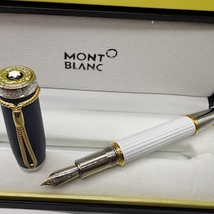 Pu&ograve; includere: Una penna stilografica Montblanc in una scatola. La penna ha un corpo bianco con dettagli dorati e un pennino argentato. Il cappuccio &egrave; blu navy con dettagli dorati. La scatola &egrave; nera e gialla con il logo Montblanc.