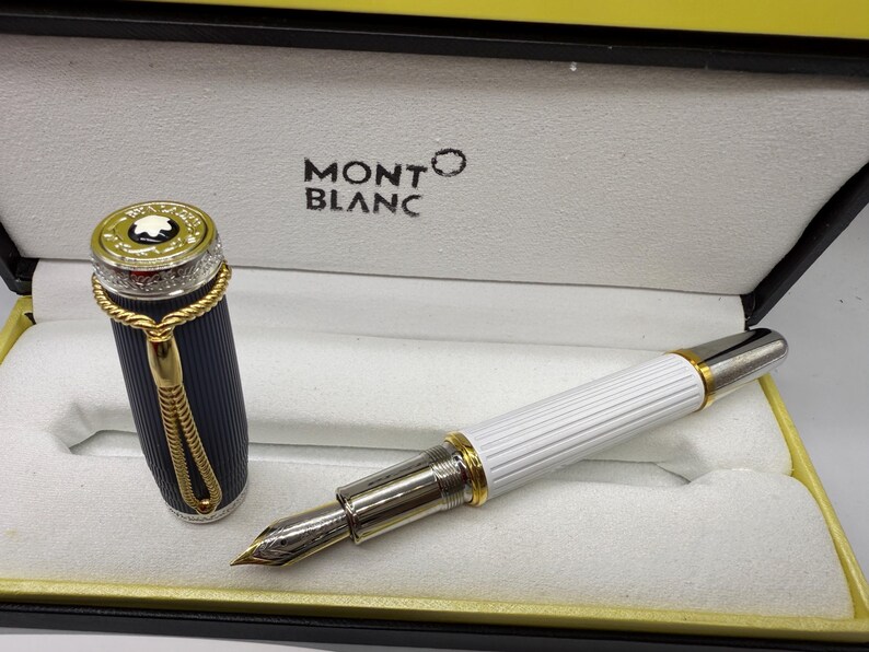 Pu&ograve; includere: Una penna stilografica Montblanc in una scatola. La penna ha un corpo bianco con dettagli dorati e un pennino argentato. Il cappuccio &egrave; blu navy con dettagli dorati. La scatola &egrave; nera e gialla con il logo Montblanc.