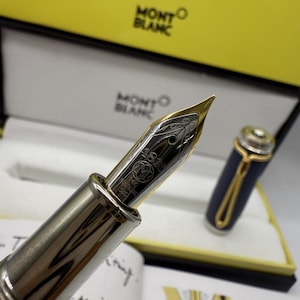 Pu&ograve; includere: Primo piano di una penna stilografica Montblanc con pennino in oro e corpo argentato. La penna &egrave; parzialmente svitata, rivelando l'intricato design del pennino. La penna &egrave; presentata in una scatola gialla e bianca con il logo Montblanc.