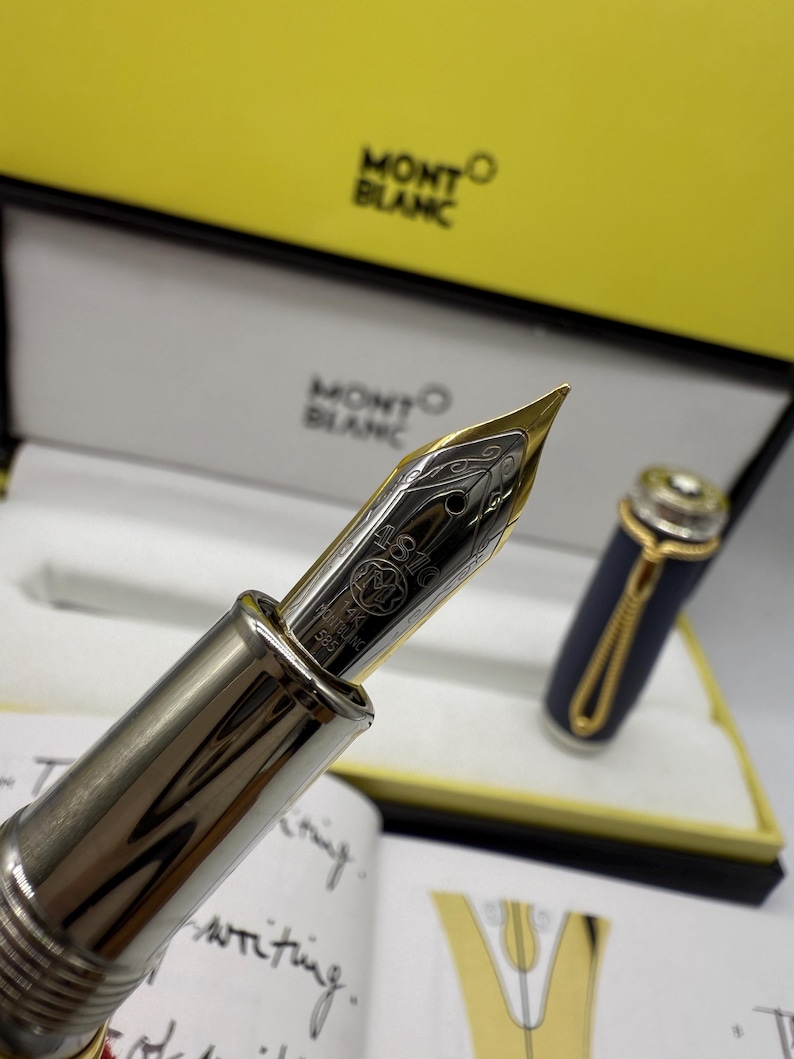 Pu&ograve; includere: Primo piano di una penna stilografica Montblanc con pennino in oro e corpo argentato. La penna &egrave; parzialmente svitata, rivelando l'intricato design del pennino. La penna &egrave; presentata in una scatola gialla e bianca con il logo Montblanc.