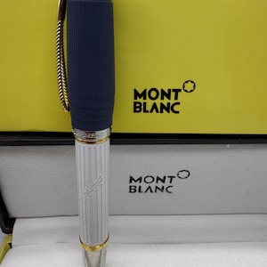 Pu&ograve; includere: Una penna Montblanc con un fusto blu navy a coste e una parte inferiore bianca con linee verticali. La penna ha dettagli color oro e una punta argentata. La penna &egrave; esposta nella sua scatola originale con il logo Montblanc.