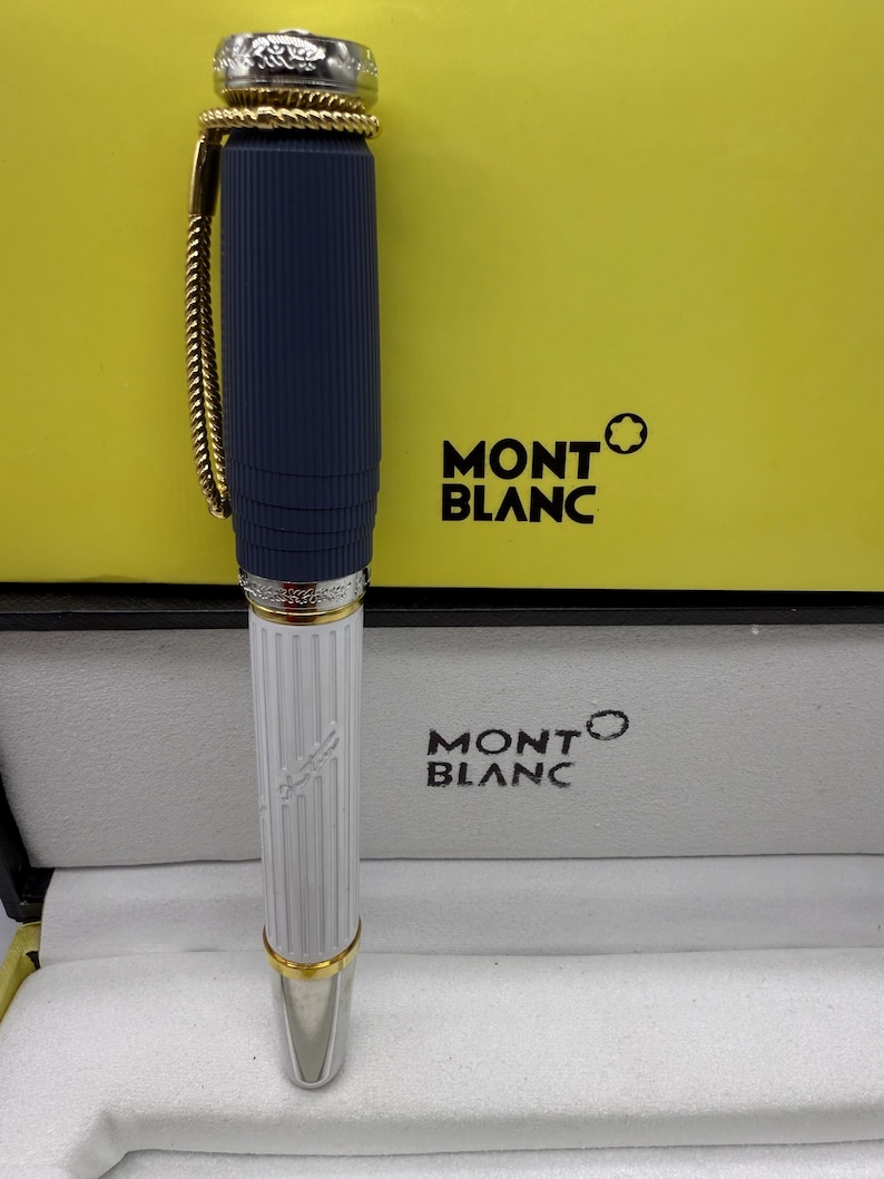 Pu&ograve; includere: Una penna Montblanc con un fusto blu navy a coste e una parte inferiore bianca con linee verticali. La penna ha dettagli color oro e una punta argentata. La penna &egrave; esposta nella sua scatola originale con il logo Montblanc.