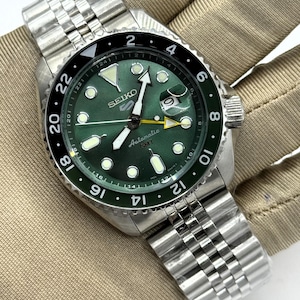 May include: A Seiko 5 Automatic GMT watch with a sunburst green dial, luminous circular indices, and a yellow GMT hand. The watch features a stainless steel jubilee-style bracelet, a date window with a cyclops lens, and a black and green bezel.