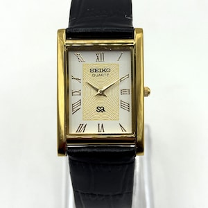 May include: A rectangular gold-toned Seiko Quartz watch with a white face and black leather band. The watch face features Roman numerals and gold hands. The watch has a gold crown and a black leather band.