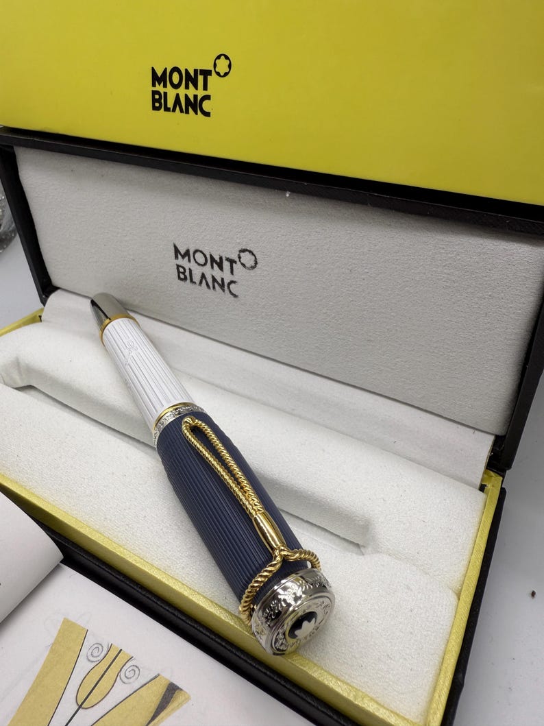 Pu&ograve; includere: Una penna Montblanc in una scatola gialla e nera. La penna ha un corpo bianco e blu navy con dettagli dorati. La penna &egrave; in una scatola foderata bianca con il logo Montblanc. La scatola &egrave; all'interno di una scatola gialla con il logo Montblanc.