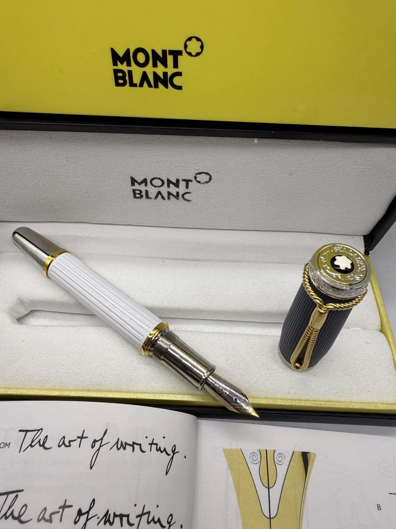 Pu&ograve; includere: Una penna stilografica Montblanc nella sua scatola di presentazione. La penna ha un fusto bianco e scanalato con dettagli dorati e argentati. Il cappuccio &egrave; staccato, rivelando un design nero e oro. La scatola &egrave; gialla e bianca con il logo Montblanc.