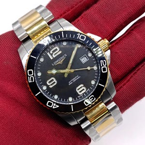 May include: A two-tone Longines automatic wristwatch featuring a black dial with gold-tone hands and markers, large Arabic numerals at 6, 9, and 12 o'clock, a date window at 3 o'clock, and a black rotating bezel on a stainless steel and gold-tone link bracelet.