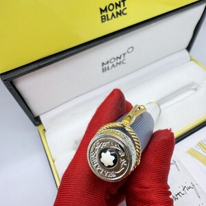 Pu&ograve; includere: Una penna Montblanc in una scatola gialla e nera. La penna ha un design argentato e dorato con un emblema in bianco e nero. La penna &egrave; tenuta da una persona con guanti rossi. La scatola ha il logo Montblanc.