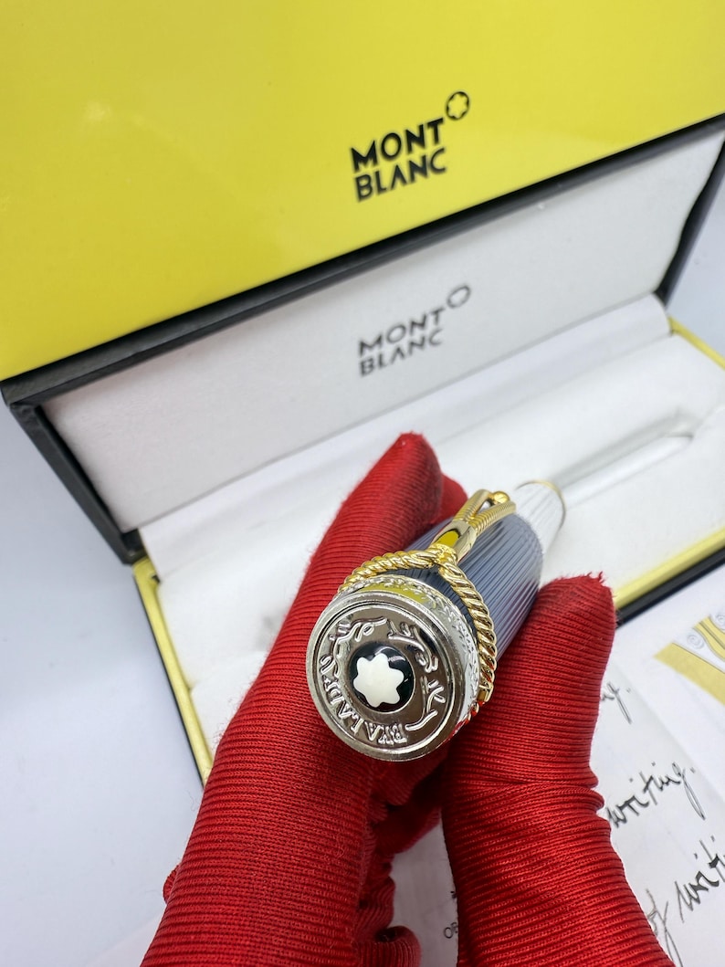 Pu&ograve; includere: Una penna Montblanc in una scatola gialla e nera. La penna ha un design argentato e dorato con un emblema in bianco e nero. La penna &egrave; tenuta da una persona con guanti rossi. La scatola ha il logo Montblanc.