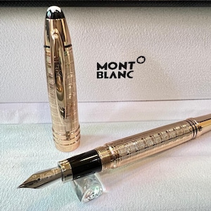 May include: A gold-colored fountain pen with a detailed engraved pattern, lying next to its cap. The pen has a black nib and a black grip section. The pen is displayed in a white box with the "MONTBLANC" logo.