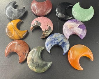 May include: A collection of polished crescent moon-shaped gemstones in various colors, including red, black, green, yellow, purple, blue, and orange. The gemstones are arranged on a dark surface.