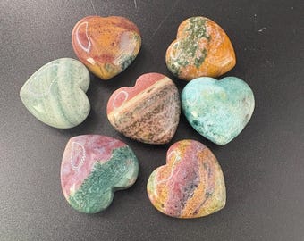 May include: Seven heart-shaped stones in various colors, including green, red, orange, and blue. The stones have a polished, smooth surface and are arranged on a dark background. The stones appear to be made of natural materials.