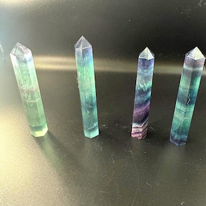 May include: Four fluorite crystal points in shades of green, blue, and purple. Each crystal has a pointed top and a rectangular base. The crystals are arranged on a dark surface. The crystals are approximately 8 cm in height.