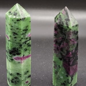 Könnte beinhalten: Zwei polierte Edelsteintürme. Die Steine sind hauptsächlich grün mit schwarzen und magentafarbenen Einschlüssen. Die Türme haben eine facettierte, spitze Spitze und eine rechteckige Basis. Die Edelsteine werden vor einem dunklen Hintergrund präsentiert.