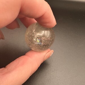 Può includere: Una sfera di cristallo trasparente, sferica, con fratture interne e inclusioni iridescenti. La sfera è tenuta tra due dita, mostrando la sua qualità traslucida e il gioco di luce all'interno del cristallo. Oggetto decorativo.