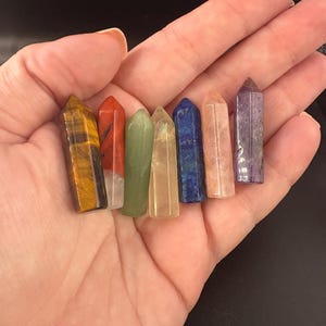 Könnte beinhalten: Eine Sammlung von sieben polierten Kristallstäben in verschiedenen Farben, darunter Gelb, Rot, Grün, Beige, Blau, Pfirsich und Lila. Die Kristalle werden in einer offenen Handfläche vor einem dunklen Hintergrund gehalten. Jeder Stab hat eine Spitze.