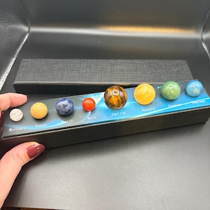 May include: A set of gemstone planets in a black box. The planets are arranged in order from Mercury to Neptune. Each planet is a different color and texture, with labels beneath each planet. The planets are held in a black box.