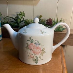 May include: White ceramic teapot with a lid and handle. The teapot is decorated with a floral design featuring pink roses and green leaves. The teapot is sitting on a wooden surface.
