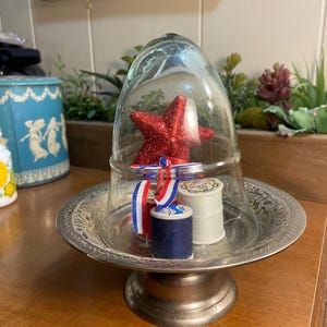May include: A decorative display featuring a red glitter star, spools of thread, and a red, white, and blue ribbon, all under a glass cloche. The display sits on a silver-colored pedestal plate.