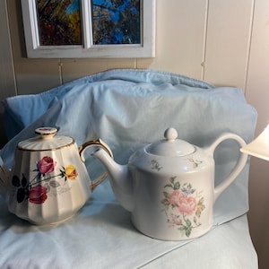 May include: Two ceramic teapots. The left teapot is cream-colored with vertical fluting, gold trim, and a floral pattern of red and yellow roses. The right teapot is white with a smooth finish and a delicate pink rose and green leaf floral design.