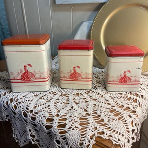 May include: Three vintage metal canisters with red and orange lids. The white canisters feature a red illustration of a woman with an umbrella. The canisters are arranged on a white lace tablecloth.