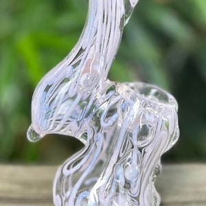 May include: A clear glass pipe featuring an upright, curved neck and a rounded base. The glass is decorated with swirling, opaque light pink and white glass ribbons that create a textured, marbled pattern throughout the entire structure.