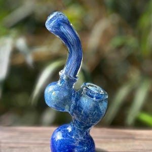 May include: A blue glass pipe with a unique shape. The pipe features a rounded base, two spherical chambers, and a curved mouthpiece. The glass has a marbled appearance with shades of blue and white. The pipe is displayed on a wooden surface.