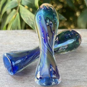 May include: Two hand-blown glass pipes with a blue, green, and gold color scheme. The pipes have a rounded shape with a flared mouthpiece and a small bowl. The glass has a swirling pattern with white accents.