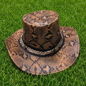 May include: A brown and black snakeskin patterned hat with a flat top and a wide brim. The hat features a black band around the base, decorated with an intricate metallic gold-toned braided trim and small circular metal accents on the sides.