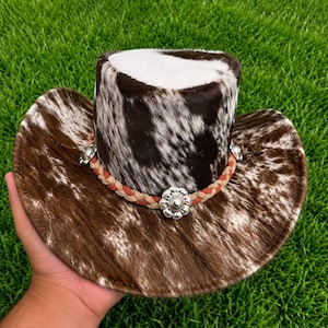 May include: A cowboy hat featuring a brown and white cowhide pattern across the crown and brim. The hat is accented with a braided tan band and silver-toned metal flower-shaped conchos spaced evenly around the base of the crown.