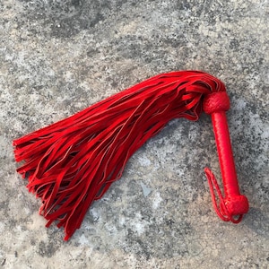 Puede incluir: Un flagelador de cuero rojo con un mango trenzado y una larga franja de tiras de cuero rojo. El mango está bien envuelto y tiene un pequeño lazo en el extremo. El flagelador está sobre una superficie gris texturizada.