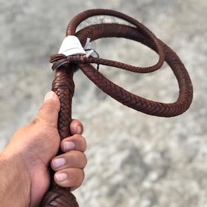 May include: A brown braided leather whip featuring a rounded knot at the base of the handle and a white wrap detail near the transition to the long, flexible lash. The whip is constructed with a consistent herringbone weave pattern throughout.