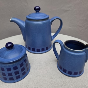 Puede incluir: Un juego de té de cerámica azul que consta de una tetera con tapa, un azucarero con tapa y una jarra de leche. Cada pieza presenta un acabado azul mate con un patrón de cuadrícula cuadrada azul oscuro en la base y pomos de tapa esféricos.