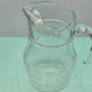 Könnte beinhalten: Ein klarer Glaskrug mit Griff und Ausguss. Der Krug hat ein strukturiertes Design um den Boden. Der Krug ist leer und steht auf einer hellblauen Oberfläche.