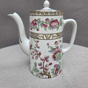 May include: A white porcelain coffee pot featuring a cylindrical body, a curved handle, and a spout. The exterior is decorated with a colourful floral pattern of pink blossoms and green leaves, accented by a gold-toned geometric border near the top.